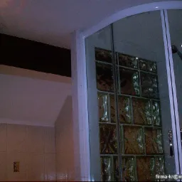 Szklana kabina prysznicowa z luksferami w łazience z beżowymi płytkami i ciemnym drewnianym elementem pod sufitem.